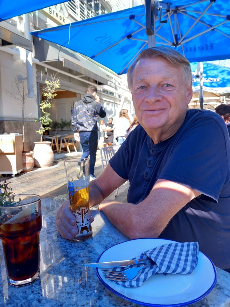 This photo shows Mark sitting at a table in the shopping precinct under a blue umbrella enjoying a cold beer