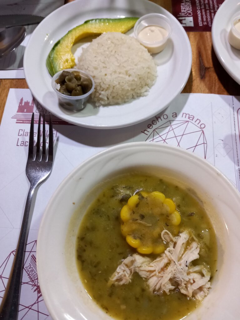 This photo shows a bowl of ajiaco with a plate of accompaniments including rice, sliced avocado, capers, and sour cream